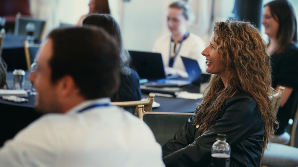 People sitting at round tables during a professional event, with laptops and notepads in front of them, smiling and engaged in conversation.
