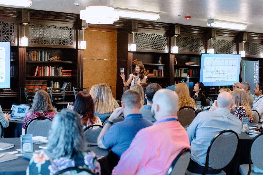A woman speaks at the front of a conference room to an audience seated at round tables, with presentation slides displayed on screens and bookshelves in the background.