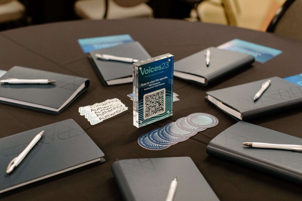 Round table set with notebooks and pens, Authenticx stickers, circular logo stickers, and a Voices23 event sign with a QR code.