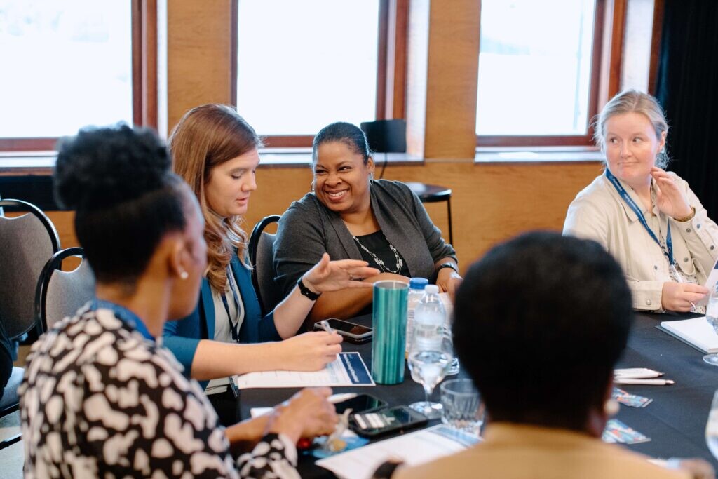 Five people sit around a conference table engaged in conversation, with notepads, water bottles, and lanyards visible, suggesting a collaborative meeting or summit event.