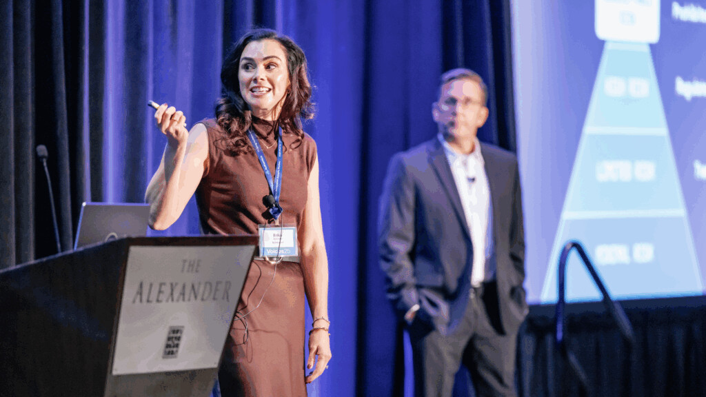 Woman speaking at a conference podium labeled The Alexander, with a man standing in the background near a projected presentation slide.